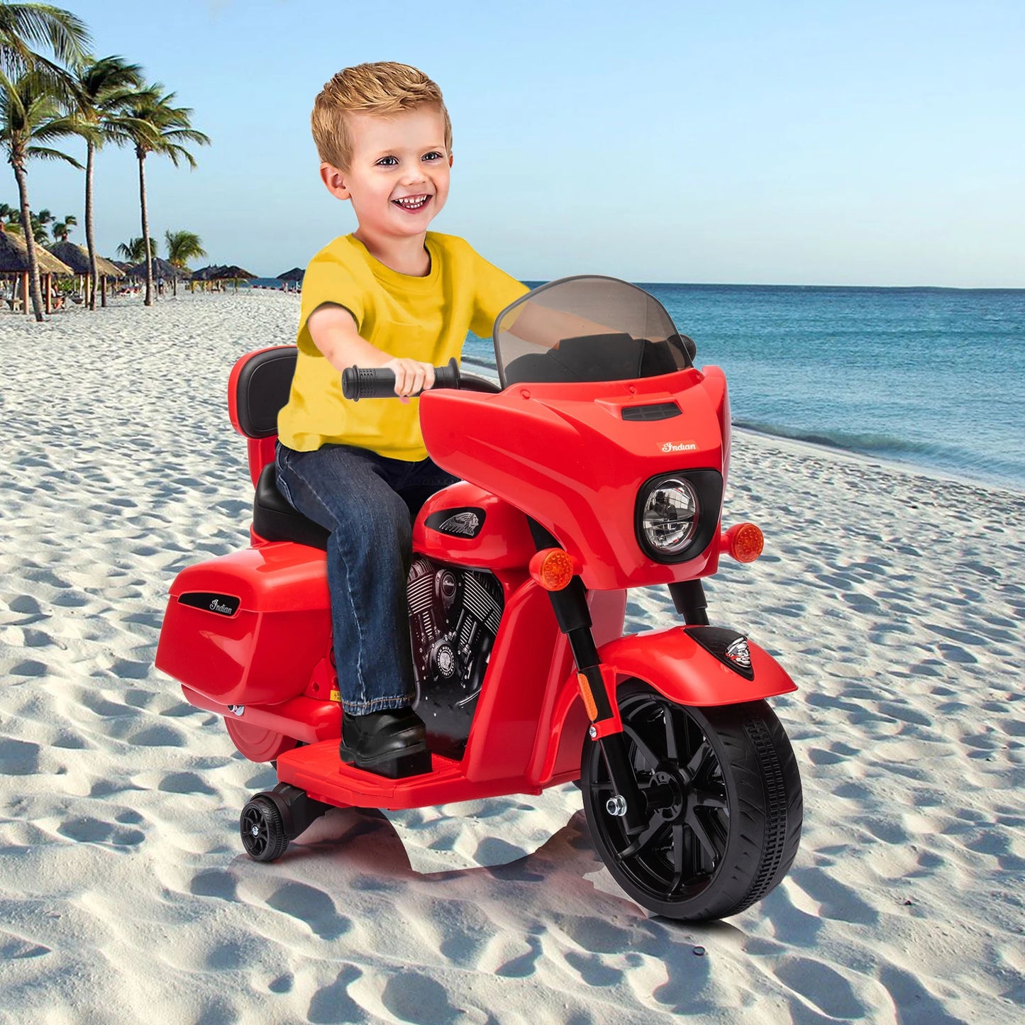 Child riding a red toy motorcycle on a sandy beach with palm trees and ocean in the background